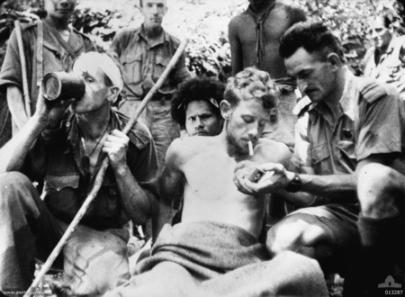 A wounded Australian soldier has his cigarette lit by Salvation Army Chaplain Albert Moore, padre to the 2/14th battalion. The soldier on the stretcher is VX51106 Lieutenant Valentine G. Gardner, D Company, 2/14th Battalion. An unidentified soldier on the left having a drink has a bandaged head and is carrying a staff. A Papuan stretcher bearer (popularly known as a Fuzzy Wuzzy Angel) is behind the stretcher. (Frame enlargement from AWM films, F01212 or F01582 taken by Damien Parer). 